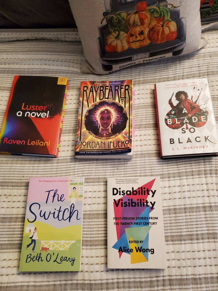 Photo of five books, in one row of three books on top and row of two books below. In the background, there is a harvest pumpkin fall pillow. The books pictured are Luster by Raven Leilani, Raybearer by Jordan Ifueko, A Blade So Black by LL McKinney, The Switch by Beth O'Leary, and Disability Visibility edited by Alice Wong.