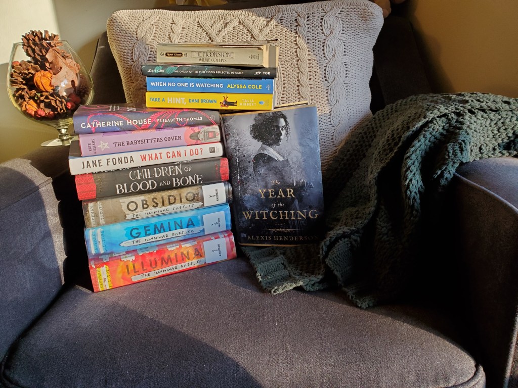 A blue armchair with a tan knit pillow and a green knit blanket. On the left arm of the chair is a large goblet with fall-ish things in (pine cones, little fake pumpkins, etc.). In the center of the photo is a stack of 11 books, with one book facing out, The Year of the Witching by Alexis Henderson.