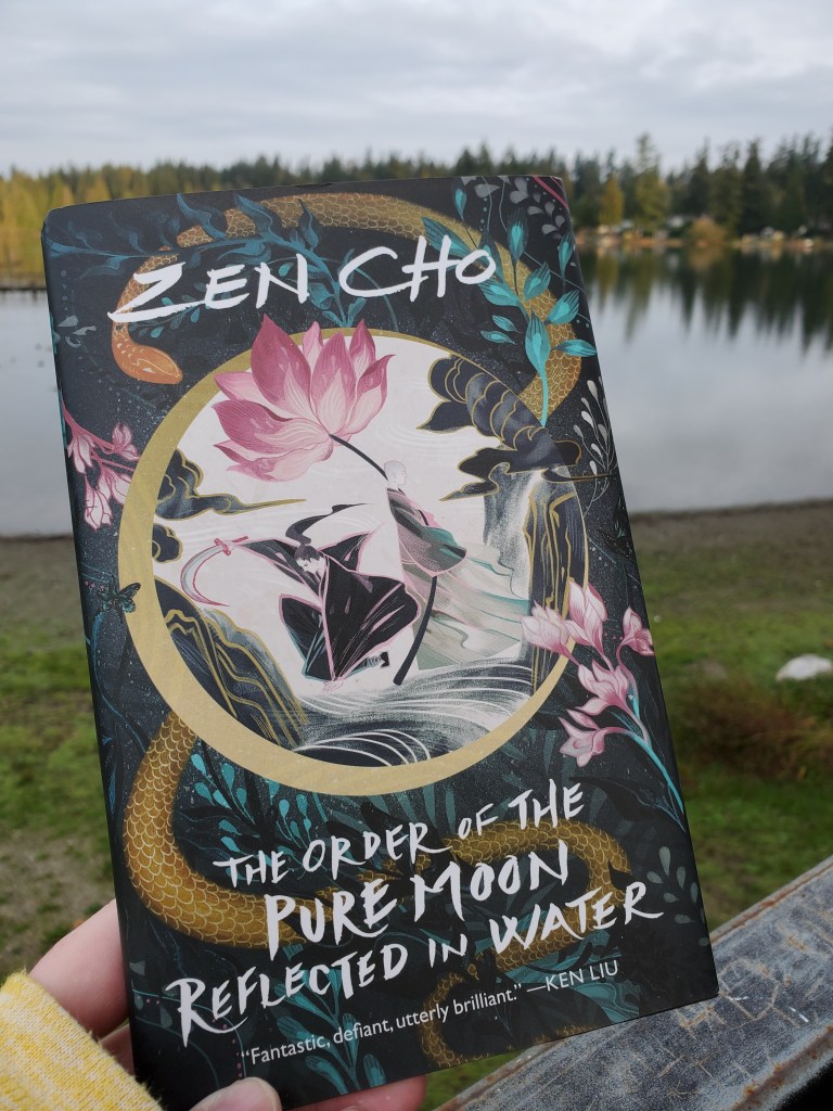 Out of focus in the background is a lake and trees. In the foreground is a copy of book The Order of the Pure Moon Reflected in Water by Zen Cho. In the center of the cover is a full moon covered by two abstract-ish figures of people, one holding a human sized long-stemmed pink flower. A snake is twined around the moon in the center, and there are smaller versions of the pink flower around the edges. There is a pull quote from author Ken Liu that says fantastic, defiant, utterly brilliant.